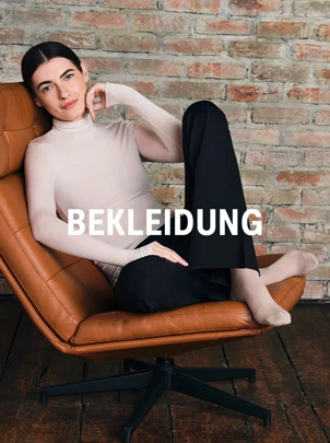 Eine Person sitzt in einem braunen Ledersessel vor einer Ziegelwand, trägt einen hellen Rollkragenpullover, schwarze Hose und helle Söckchen, darüber der Schriftzug „BEKLEIDUNG“.