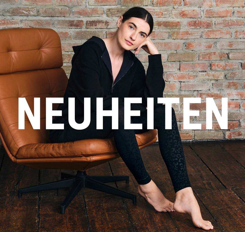 Eine Person sitzt seitlich in einem braunen Ledersessel vor einer Ziegelwand, trägt einen schwarzen Hoodie und dunkle gemusterte Leggings, mit dem groß eingeblendeten Schriftzug „NEUHEITEN“.