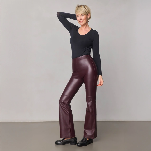 Eine Person trägt ein schwarzes Langarmshirt, bordeauxfarbene Kunstlederhose mit leichtem Schlag und schwarze Schuhe und steht vor einem einfarbig hellgrauen Hintergrund.