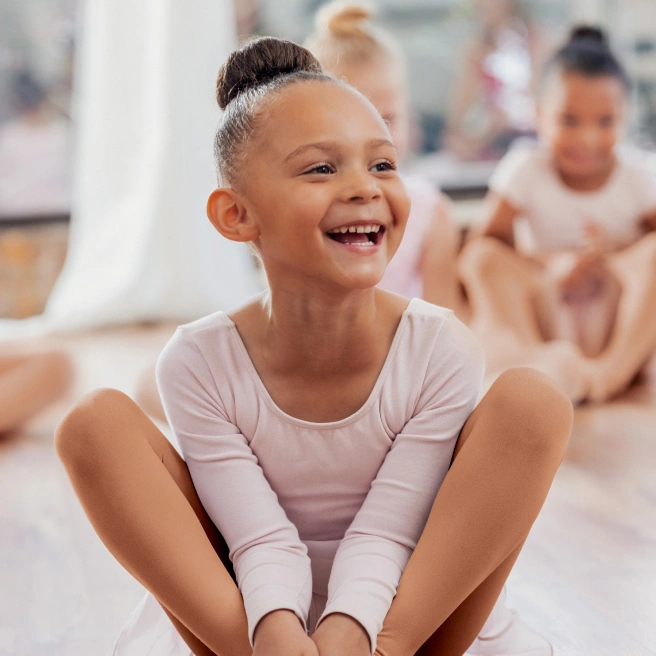 Ein Kind sitzt mit angewinkelten Beinen auf dem Boden, trägt einen hellen Langarm-Ballettanzug und hautfarbene Strumpfhosen, im Hintergrund weitere Kinder in ähnlicher Kleidung.