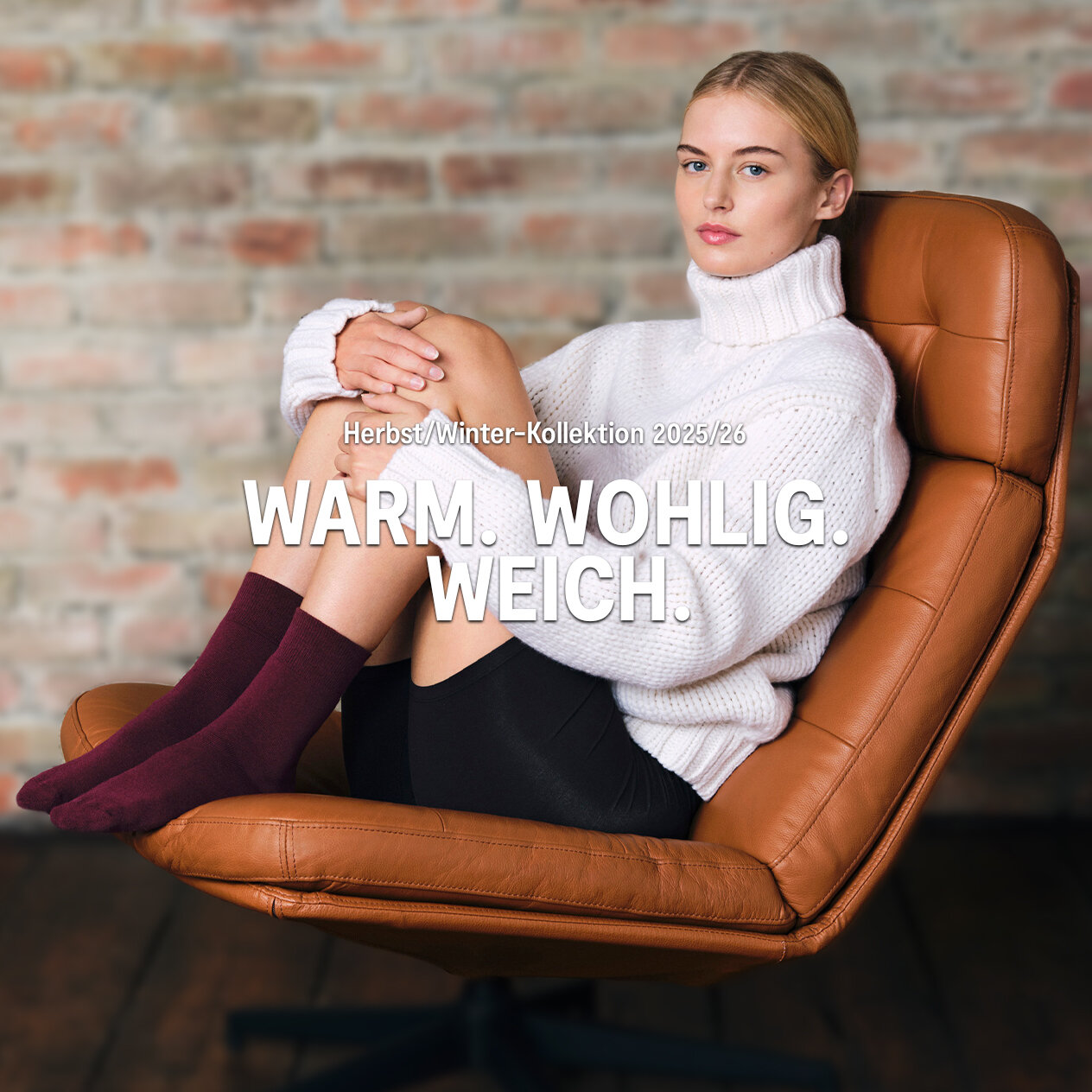 Banner für die Landingpage "Warm. Wohlig. Weich." im NUR DIE Online Shop. Eine Person in weißem Strickrollkragenpullover, schwarzen Shorts und bordeauxroten Socken sitzt mit angewinkelten Beinen in einem braunen Ledersessel vor unscharfer Ziegelwand, darüber zentriert der weiße Schriftzug „Herbst/Winter-Kollektion 2025/26 WARM. WOHLIG. WEICH.“