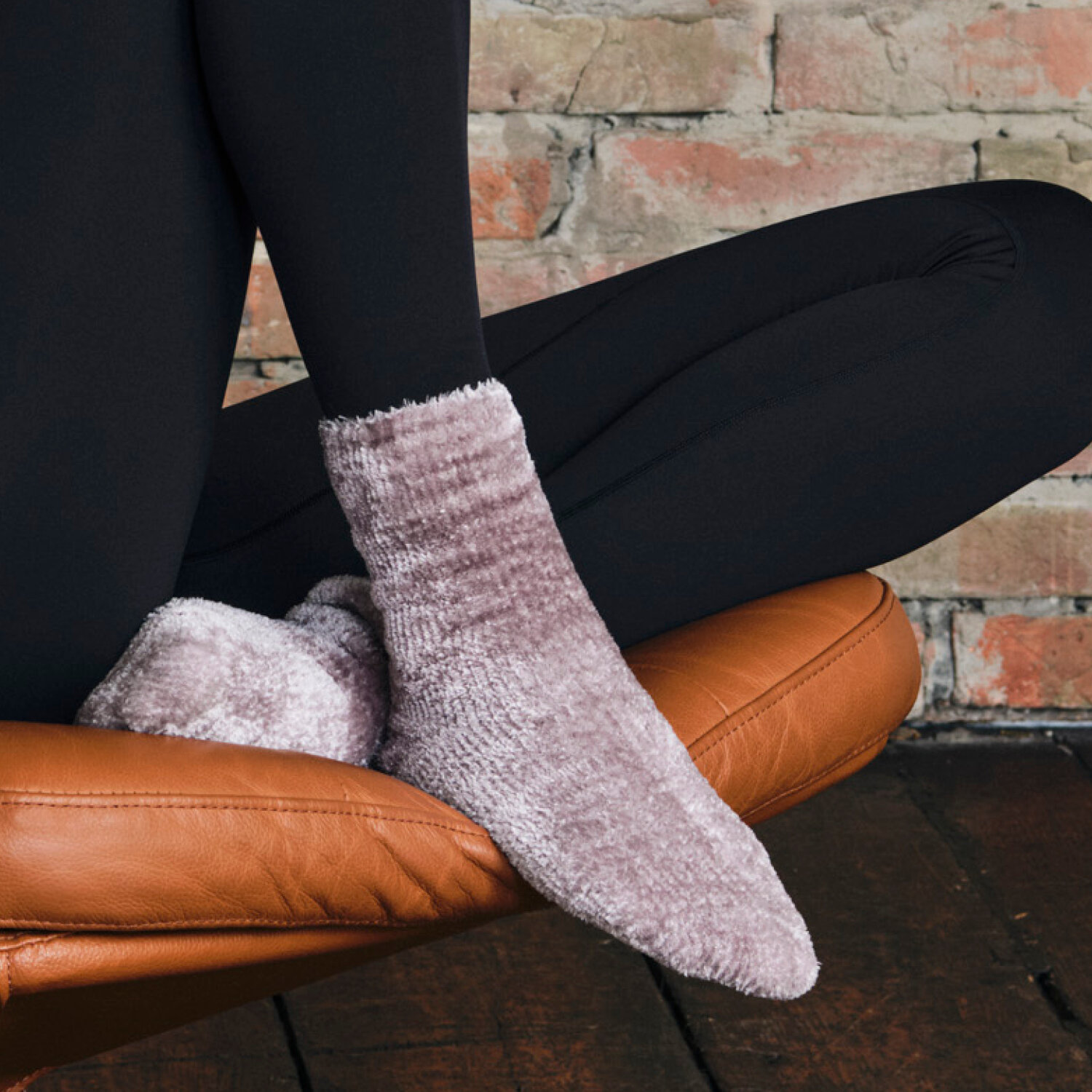 Seitliche Nahansicht von Beinen in schwarzen Leggings und flauschigen helllila-melierten Socken, angewinkelt auf einem braunen Ledersessel vor Ziegelwand und dunklem Holzboden.