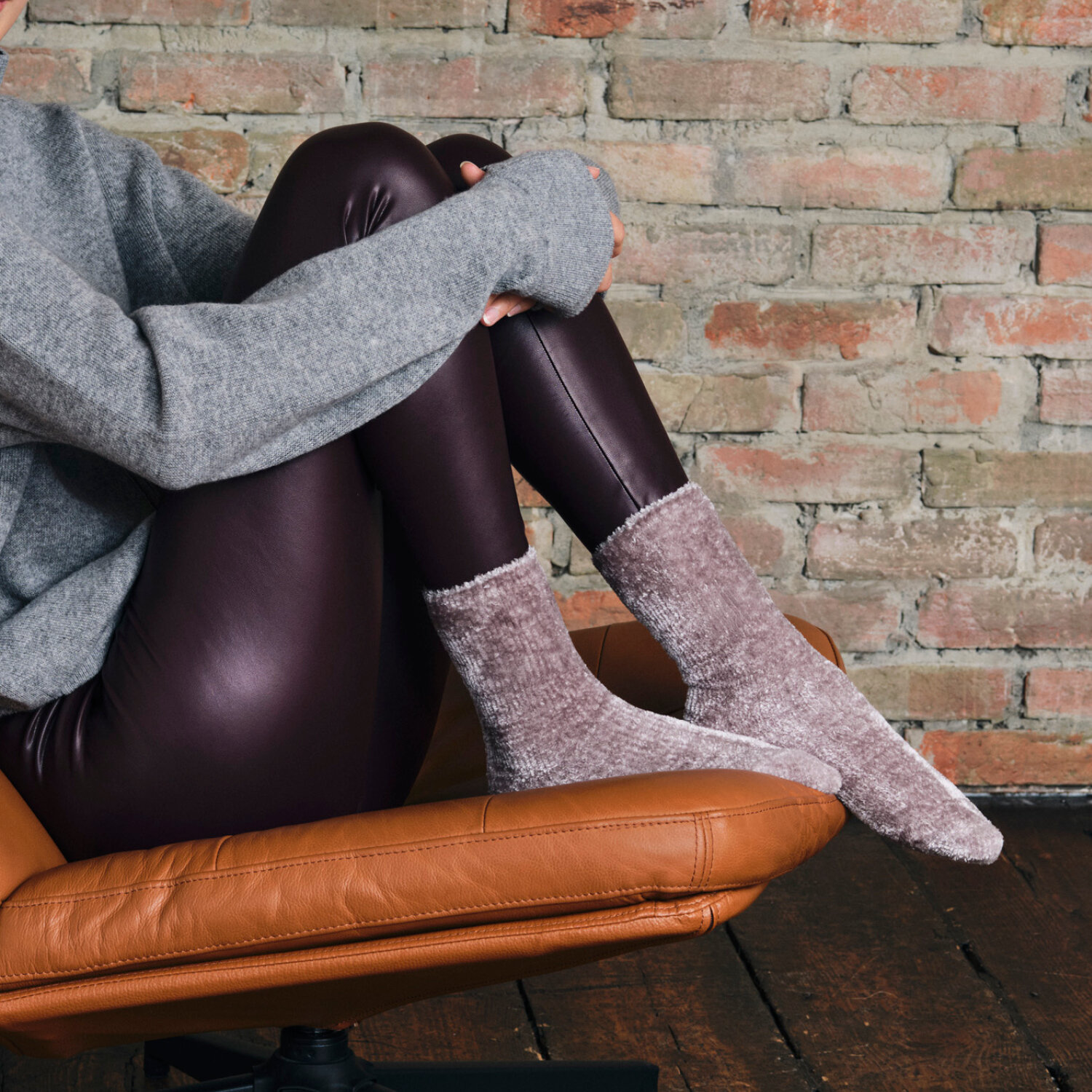 Seitliche Ansicht von Oberkörper und Beinen einer auf einem braunen Ledersessel sitzenden Person in grauem Strickoberteil, bordeauxfarbenen Lederleggings und flauschigen hellgrau-melierten Socken vor einer Ziegelwand und dunklem Holzboden.